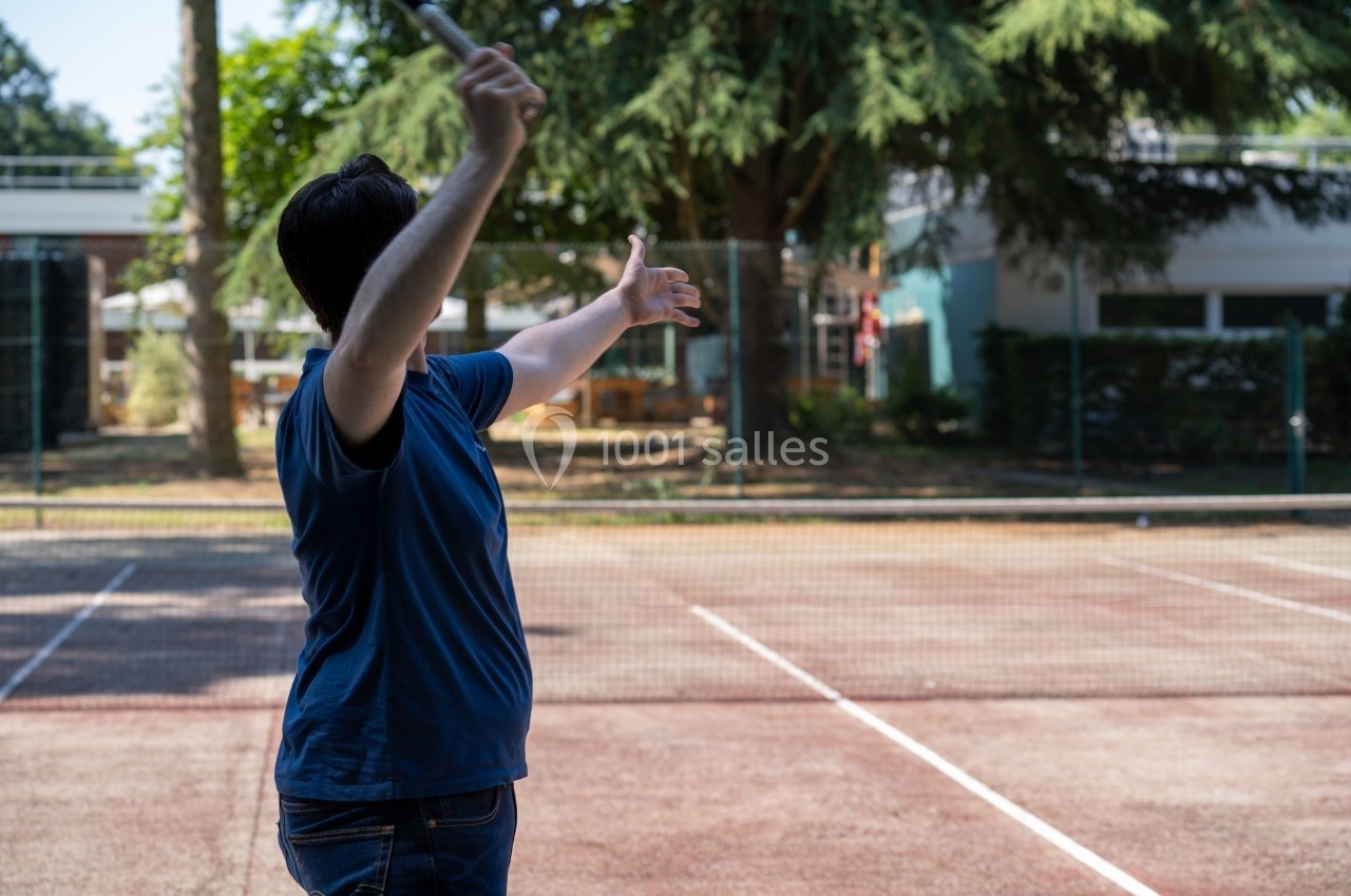 Personne en t-shirt bleu tenant une raquette, bras levés, sur un terrain de tennis extérieur ombragé.