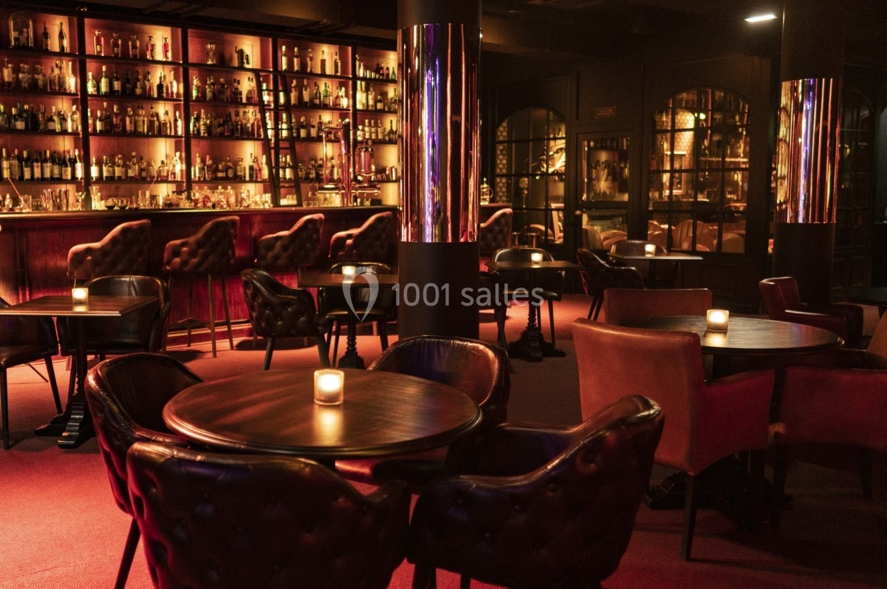Salle de bar élégante avec tables en bois, fauteuils en cuir et étagères illuminées remplies de bouteilles.