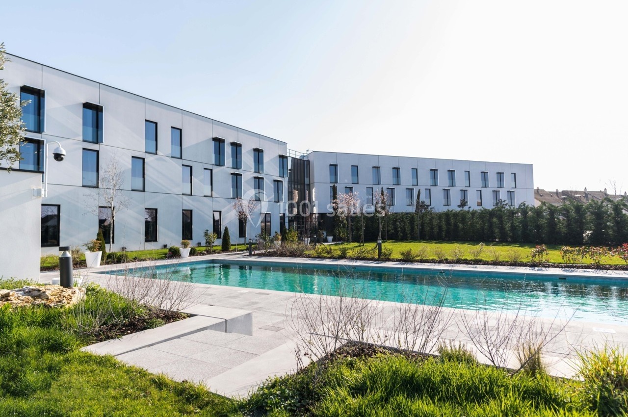 Bâtiment moderne blanc avec de grandes fenêtres, entouré d'un jardin et d'une piscine extérieure.