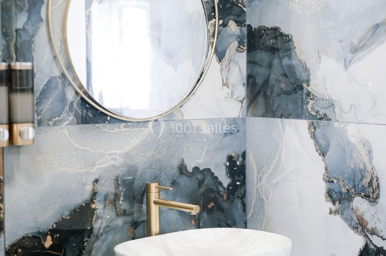 Salle de bain moderne avec un lavabo en céramique blanche, un robinet doré et un miroir rond sur un mur en marbre.