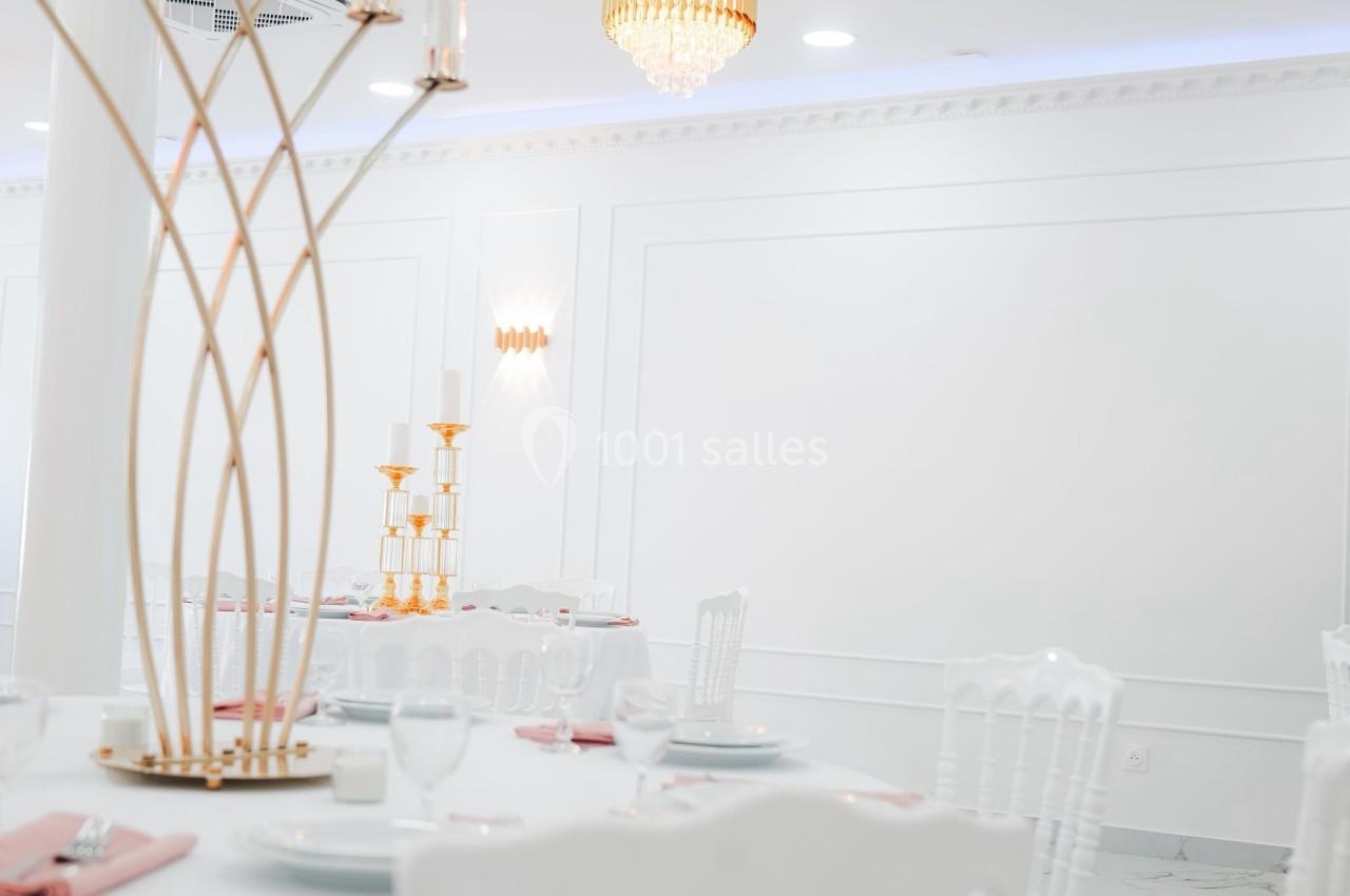 Salle de réception lumineuse avec tables dressées, chaises blanches et décorations dorées élégantes.