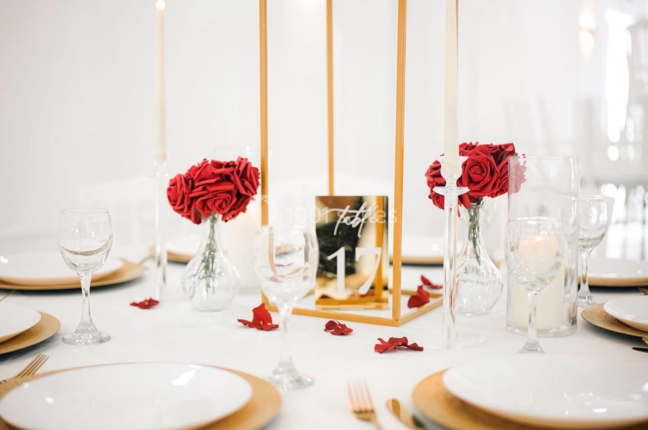 Table décorée avec des assiettes blanches, des bougies, des roses rouges et un centre de table doré.
