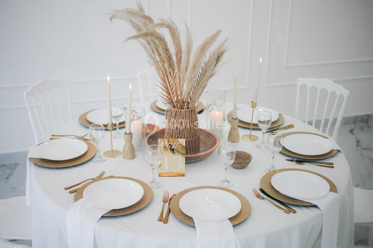 Table ronde élégamment dressée avec nappes blanches, assiettes dorées, bougies et centre de table en pampas.