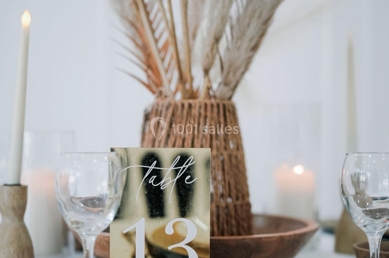 Table décorée avec des assiettes blanches, bougies, pampas et un numéro de table 13 sur un support doré.