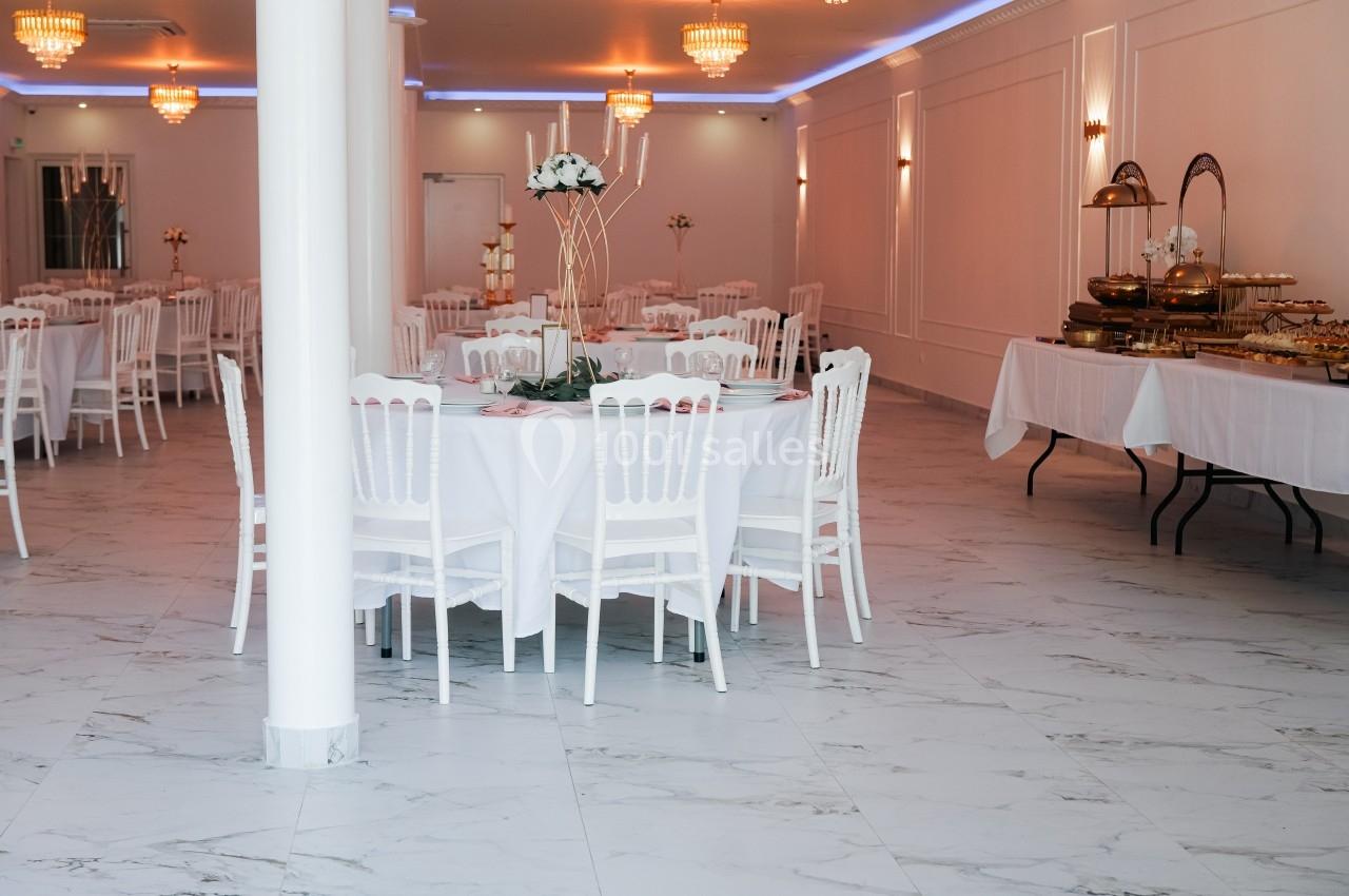 Salle de réception lumineuse avec tables rondes dressées, chaises blanches et buffets alignés sur le côté.