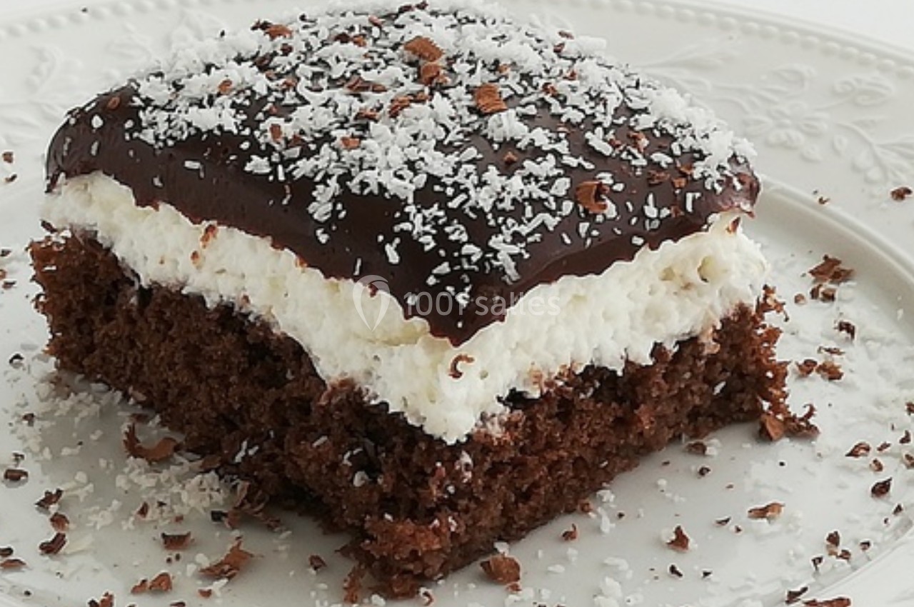 Part de gâteau au chocolat avec une couche de crème blanche, nappée de glaçage chocolat et saupoudrée de noix de coco râpée.