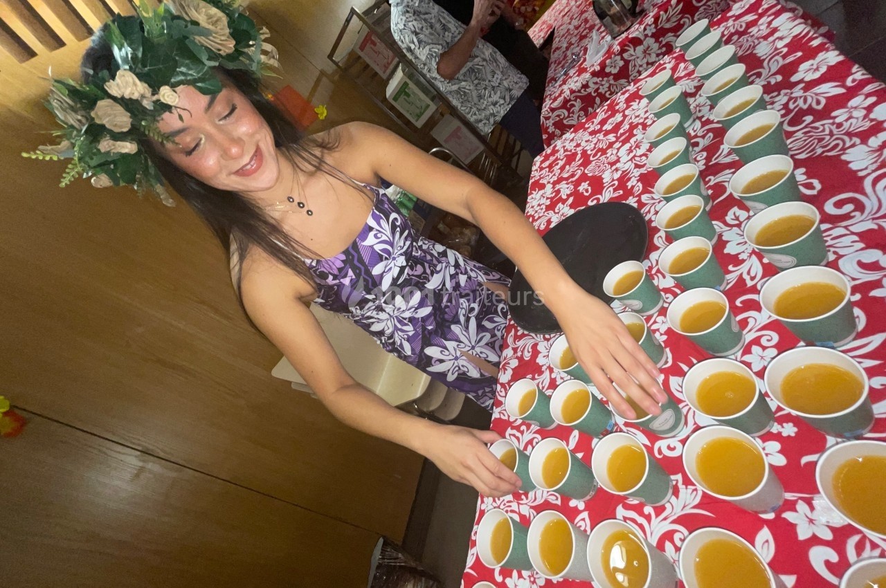 Femme en robe fleurie et couronne de feuillage disposant des tasses de boisson sur une table décorée de motifs rouges.