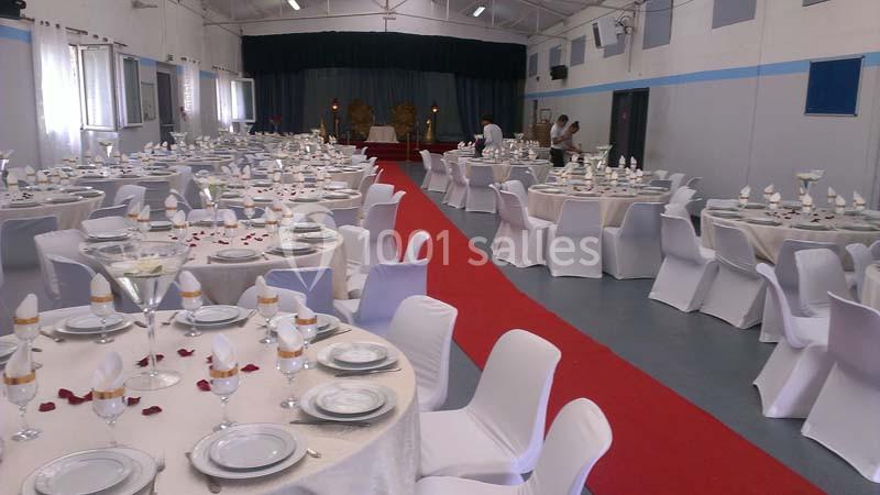 Salle de réception décorée avec des tables rondes dressées, nappes blanches et une allée centrale rouge.