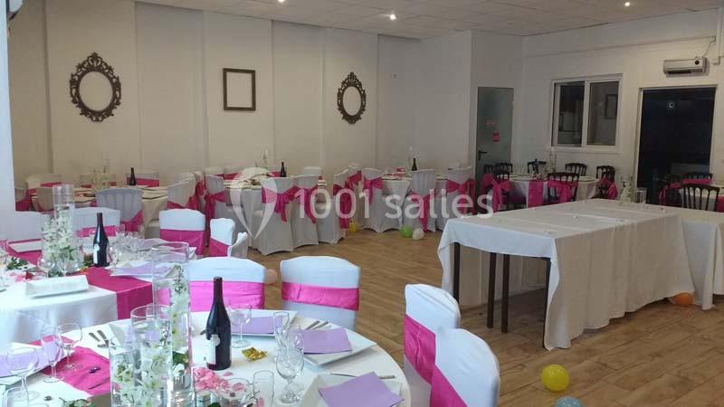 Salle décorée pour un événement avec des tables dressées, des nappes blanches et des chaises ornées de nœuds roses.