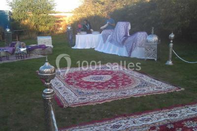 Espace extérieur décoré de tapis, lanternes et coussins, avec une table dressée en arrière-plan.