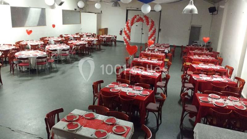 Salle décorée pour un événement, avec des tables rouges, nappes assorties et des ballons en forme de cœur.
