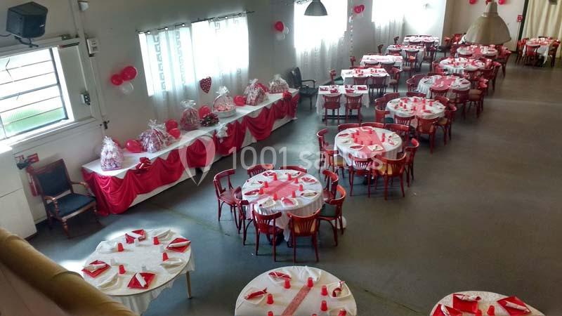 Salle décorée avec des nappes rouges et blanches, tables rondes dressées et cadeaux emballés alignés sur une table.
