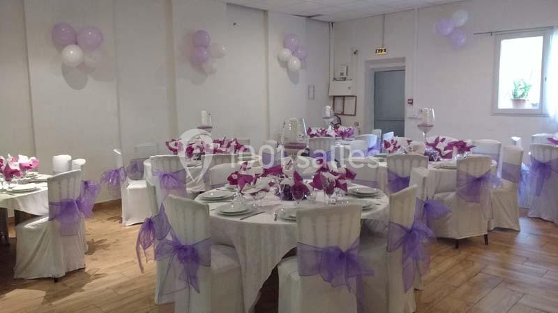 Salle décorée pour un événement avec des tables rondes, nappes blanches, chaises ornées de nœuds violets et ballons assortis.