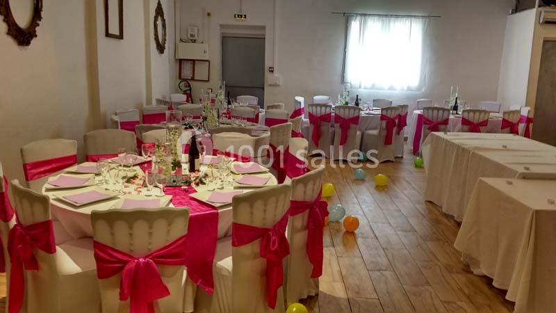 Salle décorée pour un événement avec des tables rondes dressées, nappes blanches et nœuds roses sur les chaises.