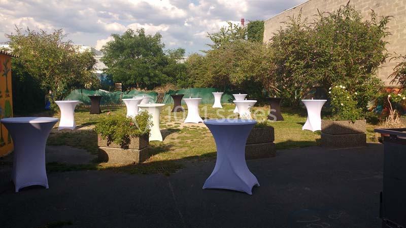 Tables hautes blanches disposées en extérieur sur une pelouse, entourées de buissons et de murs en arrière-plan.