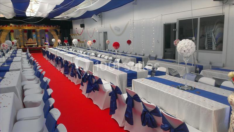 Salle décorée pour un événement avec des tables alignées, nappes blanches, nœuds bleus et tapis rouge central.