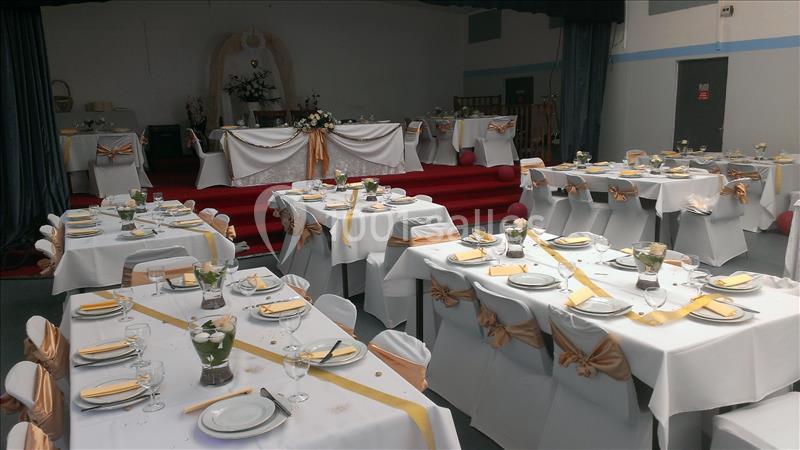 Salle décorée pour un événement avec des tables dressées, nappes blanches, chemins de table dorés et chaises ornées de…