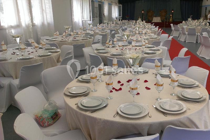 Salle de réception décorée avec des tables rondes dressées, nappes blanches, serviettes pliées et pétales rouges.
