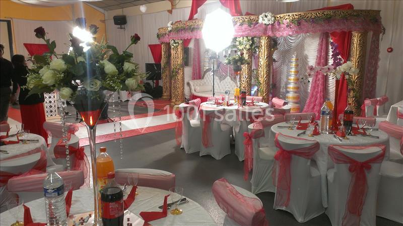 Salle décorée pour une réception avec tables ornées de nappes blanches et rubans roses, et une scène avec arche dorée.