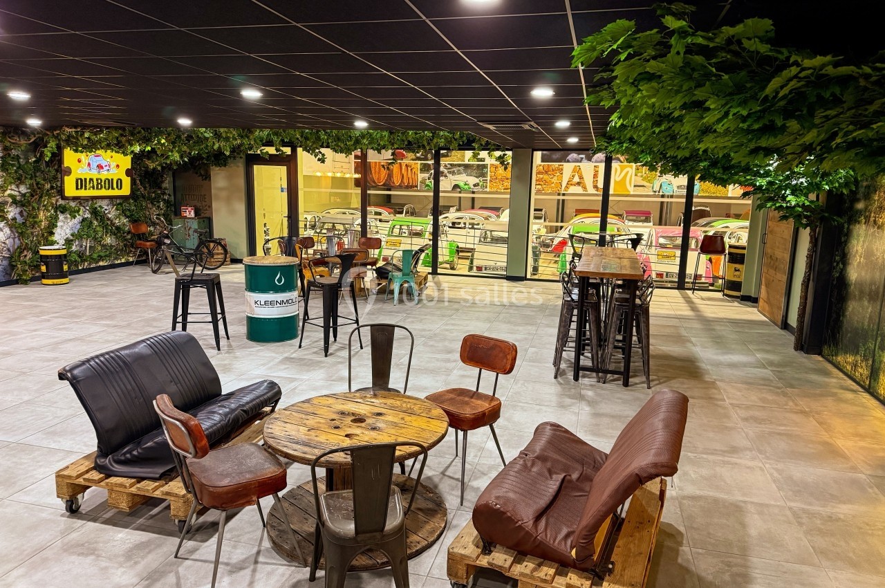 Espace intérieur avec mobilier en bois et métal, canapés sur palettes, plantes suspendues et vue sur l'extérieur vitré.
