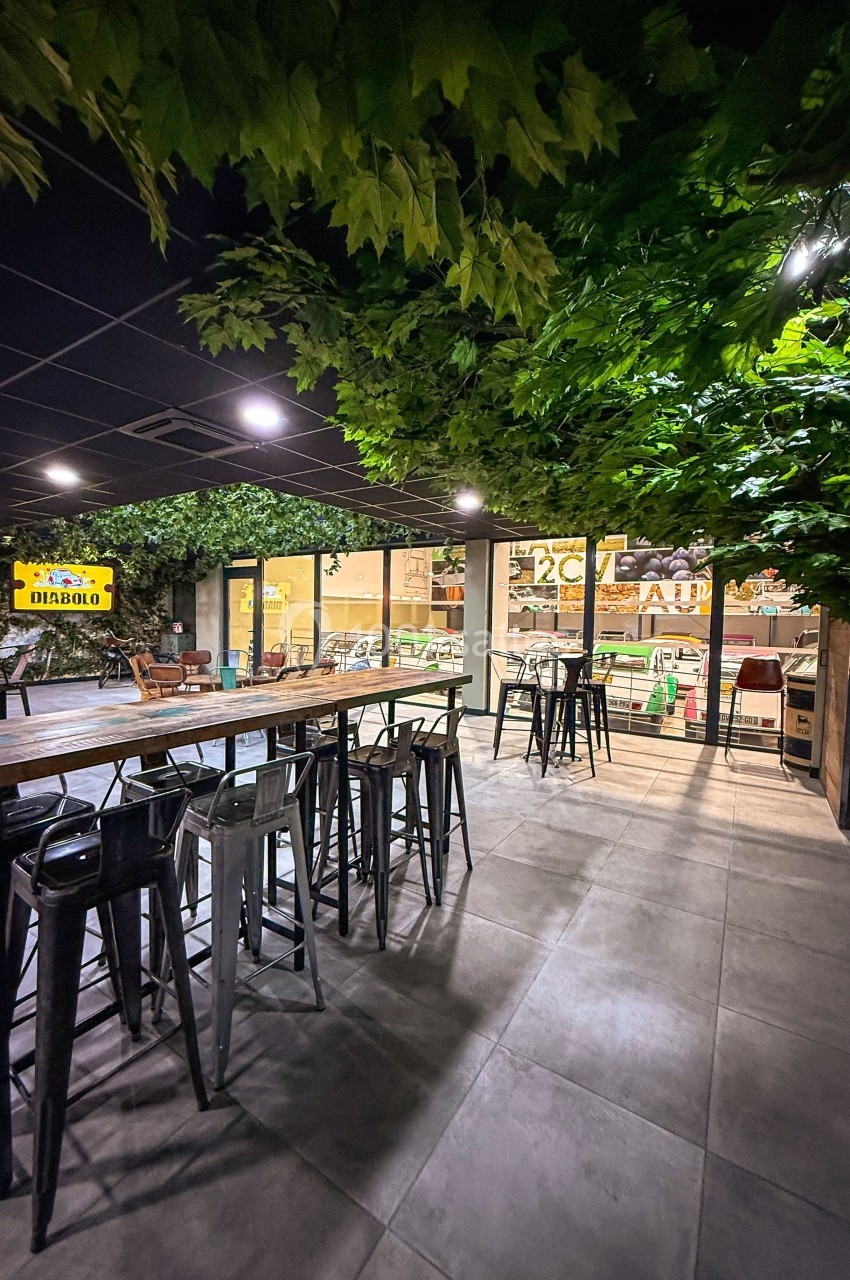 Salle intérieure avec tables hautes, tabourets noirs et plafond décoré de feuillage, donnant sur une vitrine lumineuse.