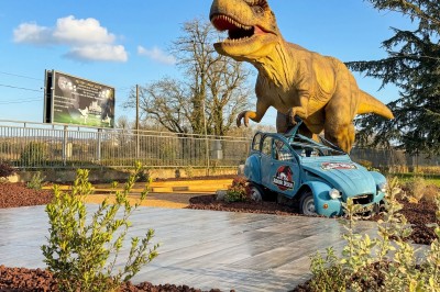 Statue d'un dinosaure attaquant une voiture bleue cabossée dans un décor extérieur avec ciel dégagé.