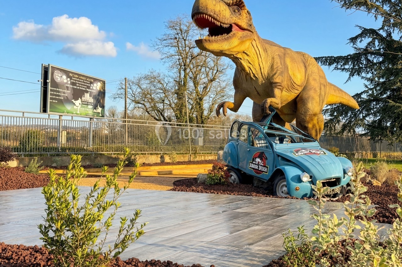 Statue d'un dinosaure T-Rex écrasant une voiture bleue dans un décor extérieur avec végétation et ciel dégagé.