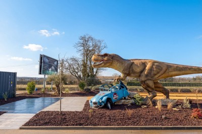 Statue d'un dinosaure attaquant une voiture bleue cabossée dans un décor extérieur avec ciel dégagé.
