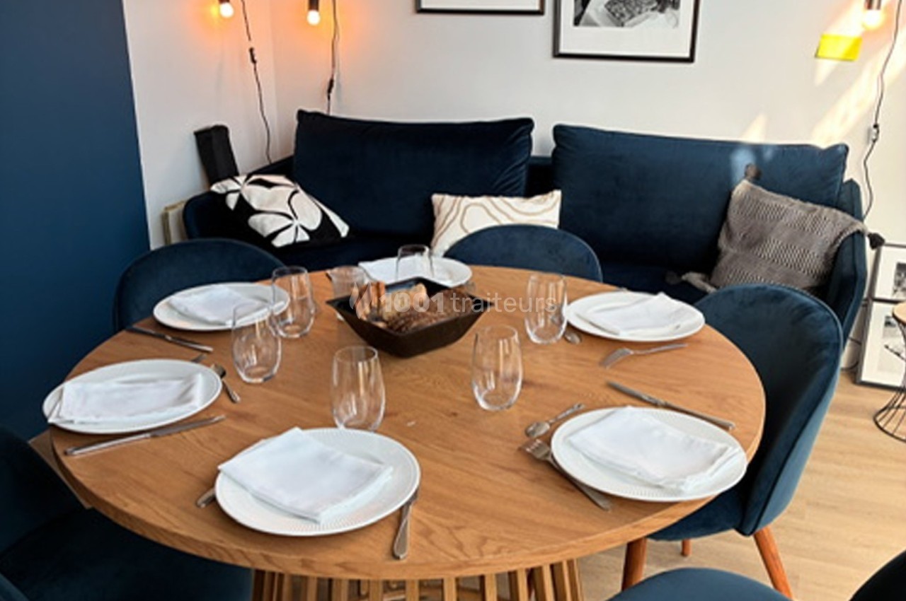 Table ronde en bois dressée avec des assiettes, verres et serviettes, entourée de chaises bleues dans un salon lumineux.