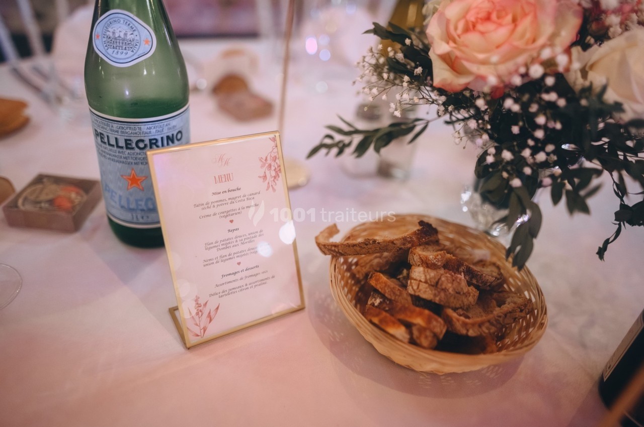 Bouteille d'eau pétillante, menu imprimé, panier de pain et bouquet de fleurs sur une table dressée.