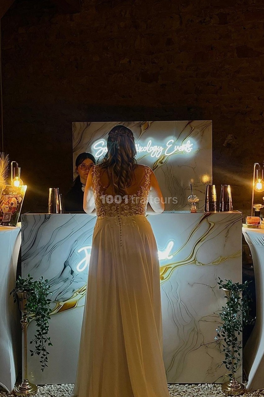 Une femme en robe blanche se tient devant un bar éclairé, décoré de marbre et de lumières chaudes.