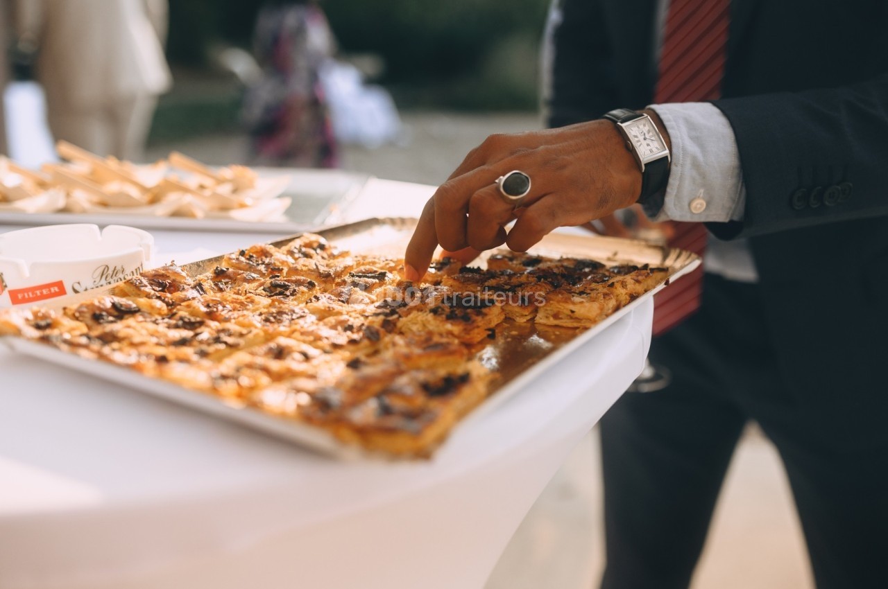 Un homme en costume prend un morceau de tarte salée sur un plateau posé sur une table blanche.