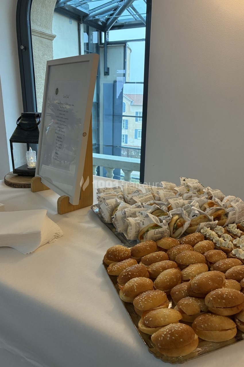 Plateau de mini-burgers et snacks emballés disposés sur une table blanche près d'une fenêtre lumineuse.
