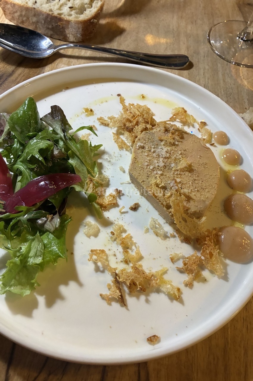 Assiette avec une tranche de foie gras, quelques gouttes de sauce, salade verte et pain sur une table en bois.