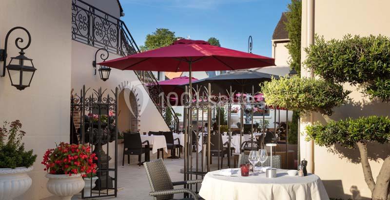 Terrasse d'un restaurant avec tables dressées, parasols rouges, plantes décoratives et architecture élégante en arrière-plan.
