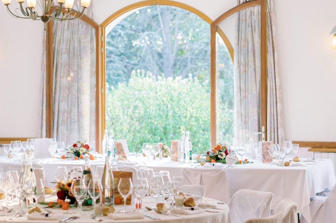 Tables élégamment dressées avec nappes blanches, verres et décorations florales, devant une grande fenêtre ouverte sur un…