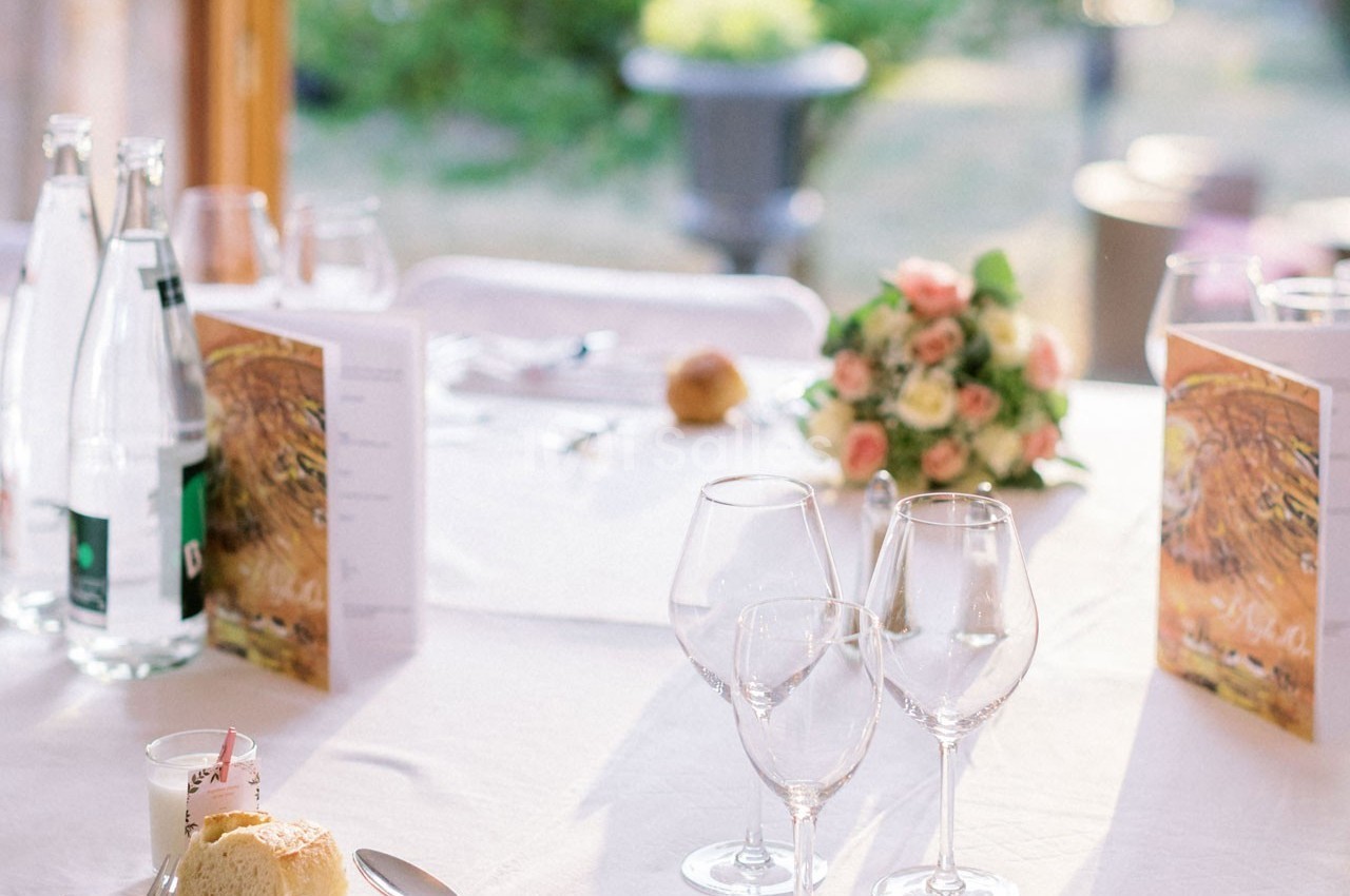 Table dressée avec nappes blanches, verres, couverts, menus, pain et bouquet de fleurs en arrière-plan flou.
