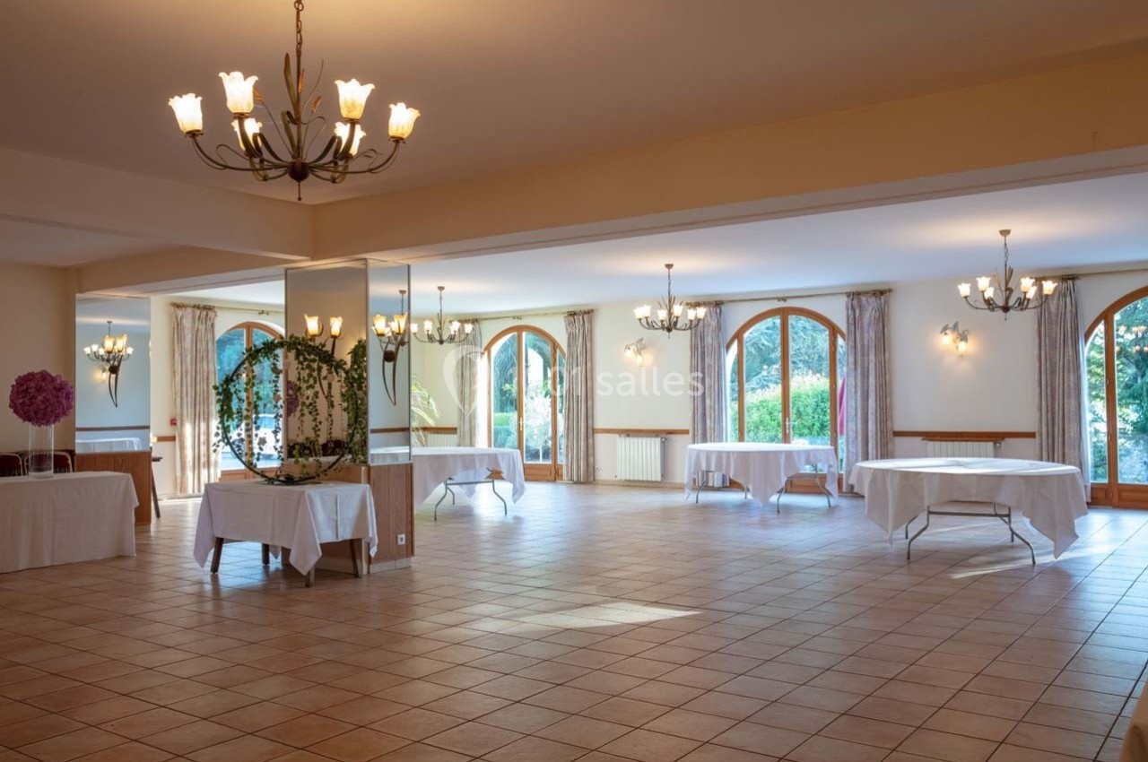 Salle lumineuse avec grandes fenêtres cintrées, tables rondes nappées blanches et lustres suspendus.