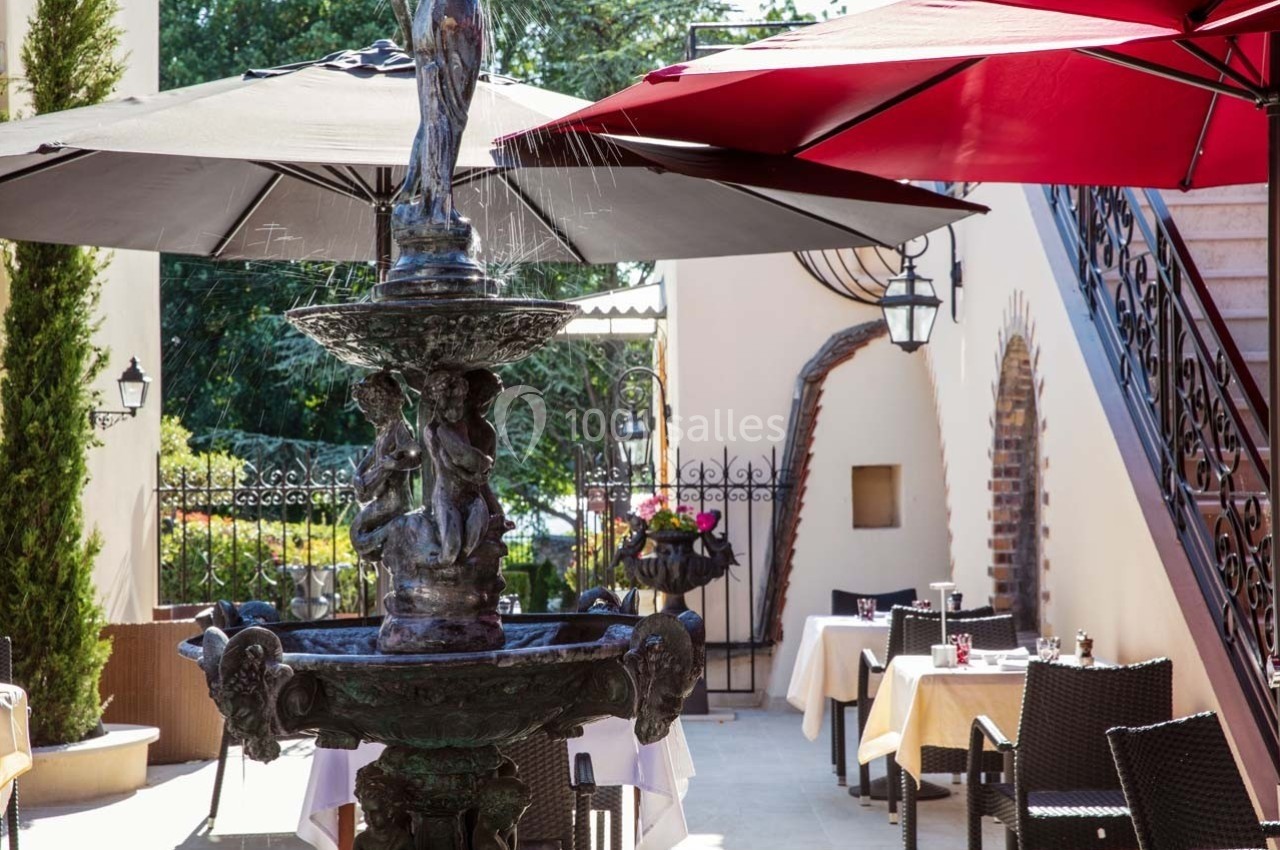 Fontaine en métal ornée de sculptures, entourée de tables dressées en terrasse avec parasols colorés.