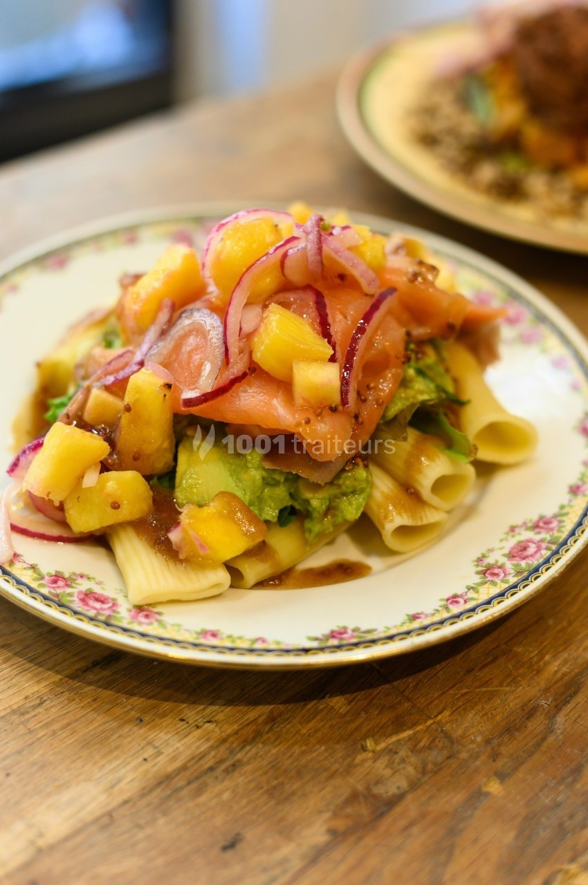 Assiette de pâtes garnie de saumon fumé, avocat, mangue, oignons rouges et vinaigrette, sur une table en bois.