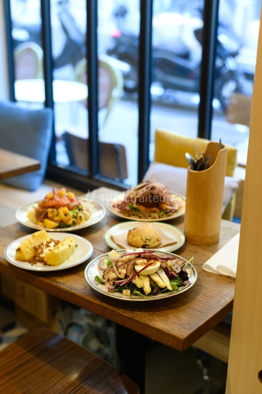 Quatre assiettes de plats variés sur une table en bois dans un restaurant avec vue sur la rue à travers une baie vitrée.
