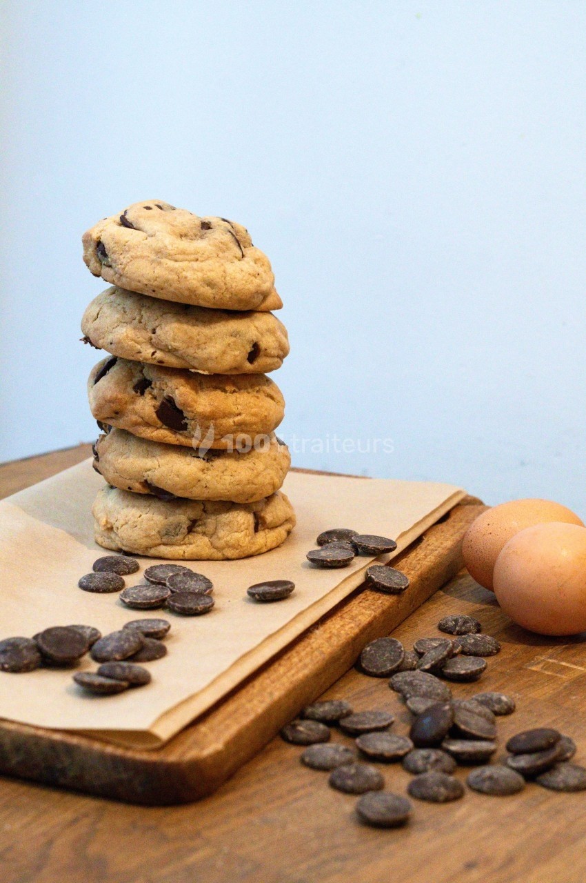Pile de cookies aux pépites de chocolat sur une planche en bois, entourée de pépites et de deux œufs.