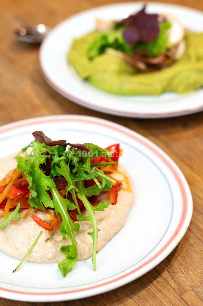 Assiette de purée garnie de légumes grillés et de salade verte, avec un autre plat flou en arrière-plan.