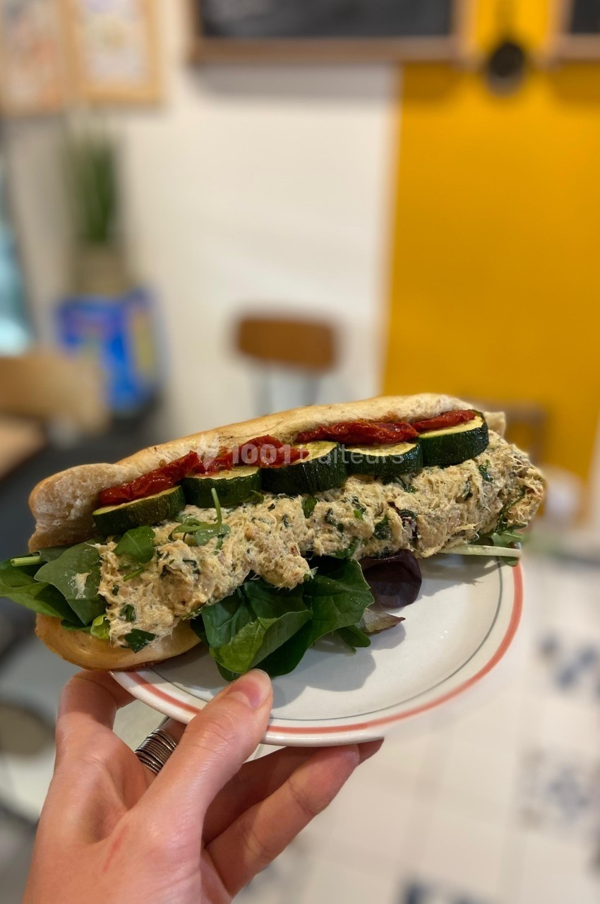 Sandwich garni de légumes grillés, feuilles de salade et tartinade, tenu dans une main dans un café lumineux.