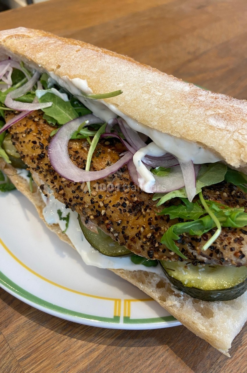 Sandwich garni de poisson grillé, légumes frais, cornichons, oignons rouges et sauce, servi sur une assiette.