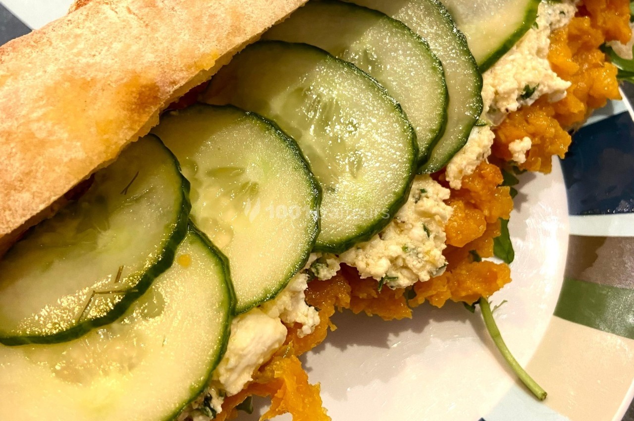 Sandwich garni de rondelles de concombre, purée de légumes orange, fromage frais et roquette sur une assiette colorée.