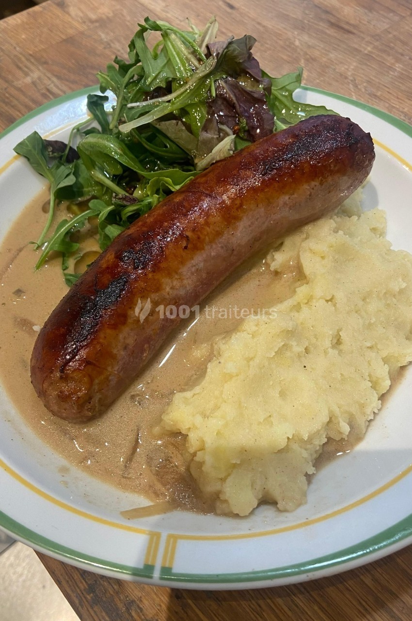 Assiette contenant une saucisse grillée, purée de pommes de terre, sauce brune et salade verte sur une table en bois.
