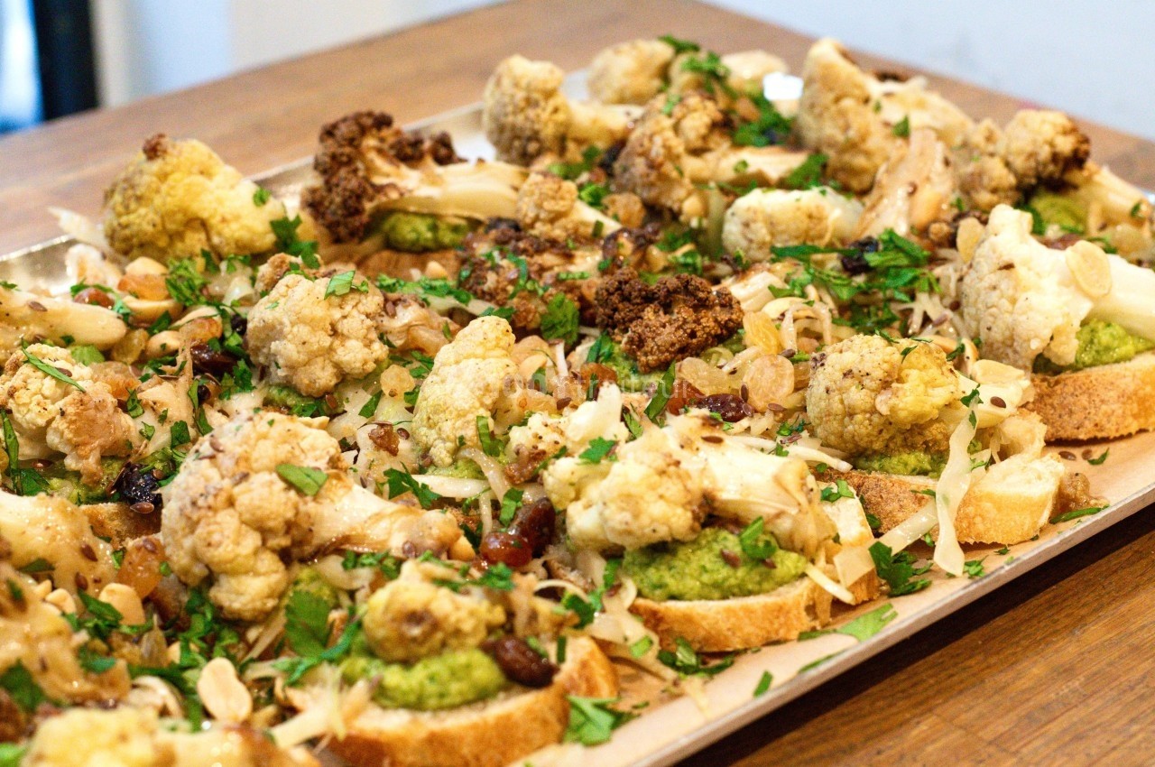 Tranches de pain garnies de chou-fleur rôti, pesto, amandes effilées et persil, disposées sur un plateau en bois.