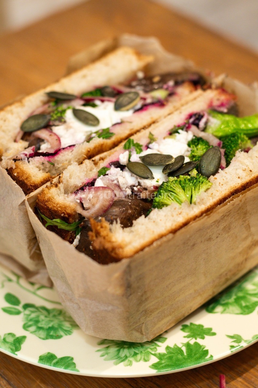 Sandwich végétarien garni de légumes, fromage frais, graines de courge et herbes, présenté sur une assiette fleurie.