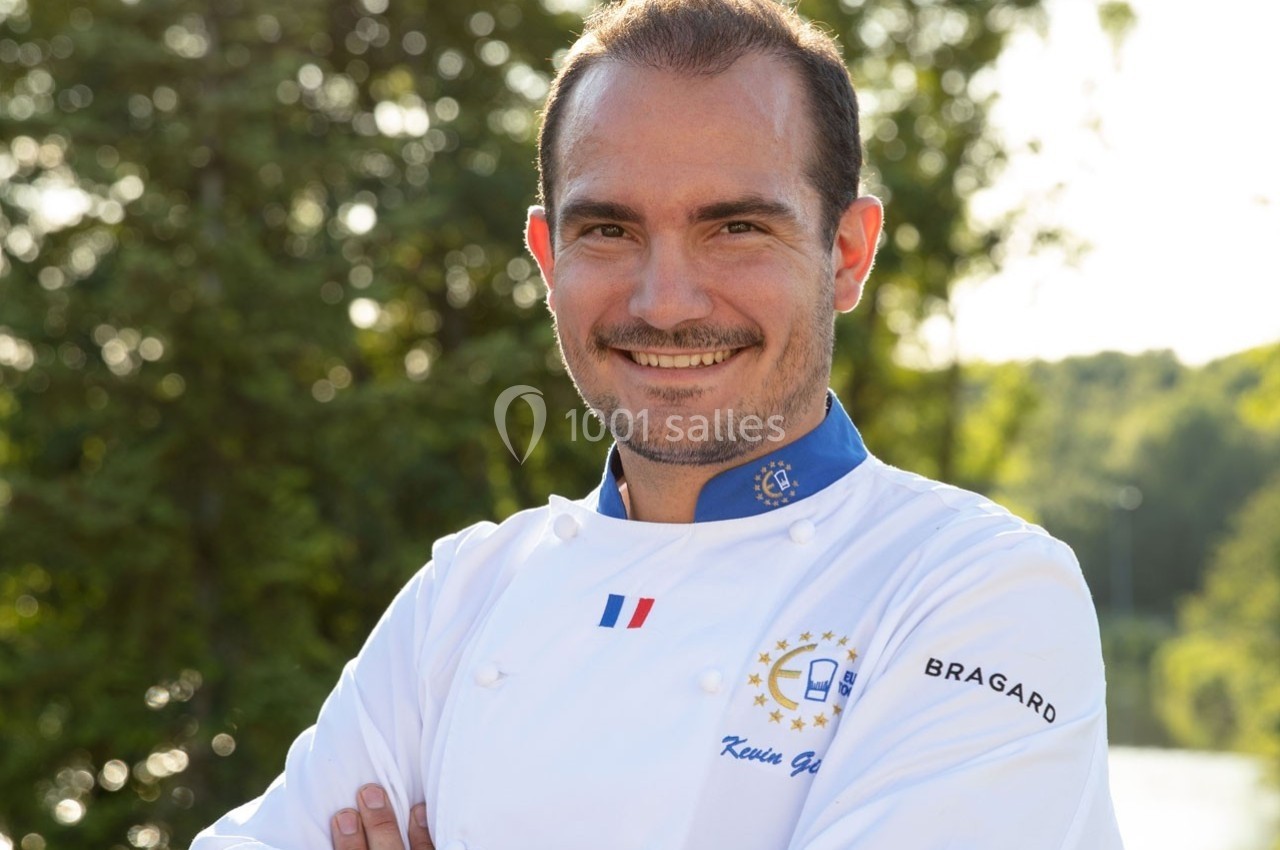 Un chef cuisinier souriant en veste blanche, debout en extérieur devant un fond d'arbres verdoyants.